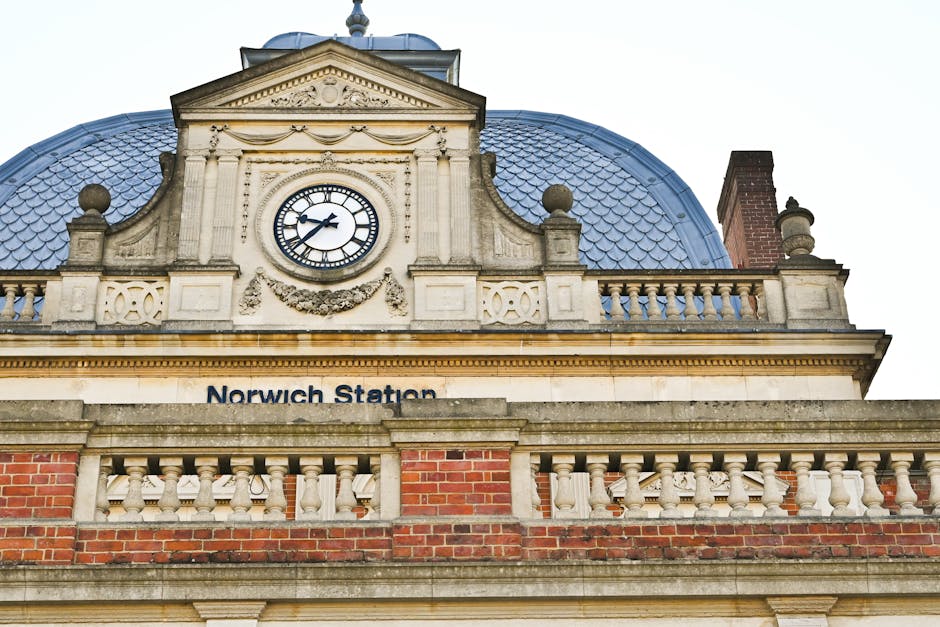 Close-up view of the upper façade of Norwich Station, featuring a decorative clock with black Roman numerals and hands, set within an ornate stone frame. The building's architecture includes classical design elements such as carved stone details, a tiled dome roof with a blue hue, and decorative balustrades made of stone and brickwork. The clock's face is clean and clearly visible, reflecting well-maintained surfaces. Part of a brick chimney can be seen on the right side, and bright natural light illuminates the scene, highlighting the details of the stonework and roof tiles. This historic station building showcases intricate architectural features and a well-presersved appearance, relevant to surface cleaning and maintenance services by Holland Park Carpet Cleaning for optimal appearance and hygiene.