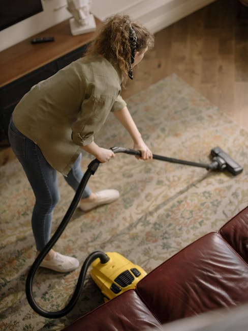 A woman with curly hair wearing a beige jacket, dark blue jeans, and white shoes is vacuuming a large, ornate area rug in a living room. She holds a yellow and black vacuum cleaner with a hose attachment, moving it across the patterned carpet which appears clean and well-maintained. The room features a wooden floor visible around the rug, with a dark brown leather sofa partially in view, and a wooden console table against the wall. The lighting is soft and natural, highlighting the tidy, polished surfaces, indicative of professional deep cleaning practices by Holland Park Carpet Cleaning for surface sanitisation and domestic cleaning maintenance.
