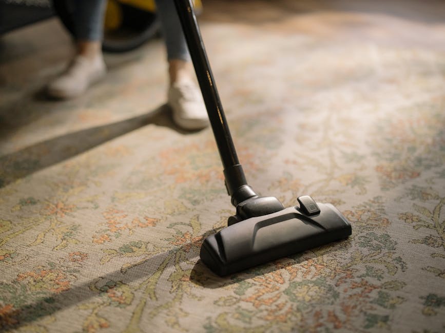 A close-up view of a person vacuuming a patterned area rug with a black vacuum cleaner attachment, positioned on a wooden floor in a residential living room. The person's feet are visible, wearing white shoes and dark pants. The room is well-lit, with natural light highlighting the clean, dust-free surface of the rug. The setting emphasizes surface cleaning and maintenance, aligning with Holland Park Carpet Cleaning's domestic cleaning services as seen in the Holland Park Avenue carpet cleaning guide for W11 homes, HOLLAND PARK.