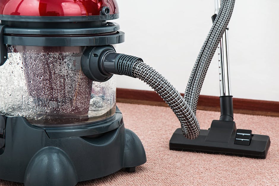 A close-up view of a person vacuuming a patterned area rug with a black vacuum cleaner attachment, positioned on a wooden floor in a residential living room. The person's feet are visible, wearing white shoes and dark pants. The room is well-lit, with natural light highlighting the clean, dust-free surface of the rug. The setting emphasizes surface cleaning and maintenance, aligning with Holland Park Carpet Cleaning's domestic cleaning services as seen in the Holland Park Avenue carpet cleaning guide for W11 homes, HOLLAND PARK.