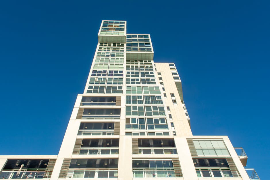 A modern high-rise building with multiple floors featuring large glass windows and white exterior panels, set against a clear blue sky. The building's surfaces appear clean and well-maintained, with no visible dust or dirt. Some windows reflect sunlight, indicating bright and natural lighting. There are balconies with glass railings and visible interior details through the windows. The image emphasizes the building's sleek architectural design and pristine appearance, aligning with surface cleaning and maintenance themes relevant to residential and commercial property care, as promoted by Holland Park Carpet Cleaning on their website for end of tenancy cleaning services in Ilchester Place, Holland Park.