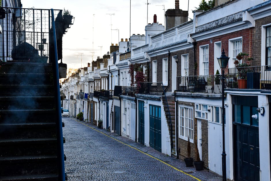 A row of residential terraced houses on Ilchester Place, showcasing brick and painted facades with white ornamental cornices and window frames. Each property has a small front balcony with black metal railings, some decorated with potted plants and outdoor furniture. The street is paved with cobblestones, and the scene is illuminated by natural daylight, with a soft, cloudy sky overhead. Visible are street lamps and chimneys on the rooftops, with a staircase on the left side leading upstairs to a building. The overall appearance is clean and well-maintained, reflecting a typical London residential area in Holland Park, which is suitable for professional domestic cleaning services. Holland Park Carpet Cleaning offers comprehensive surface cleaning and sanitisation to keep such properties in pristine condition.