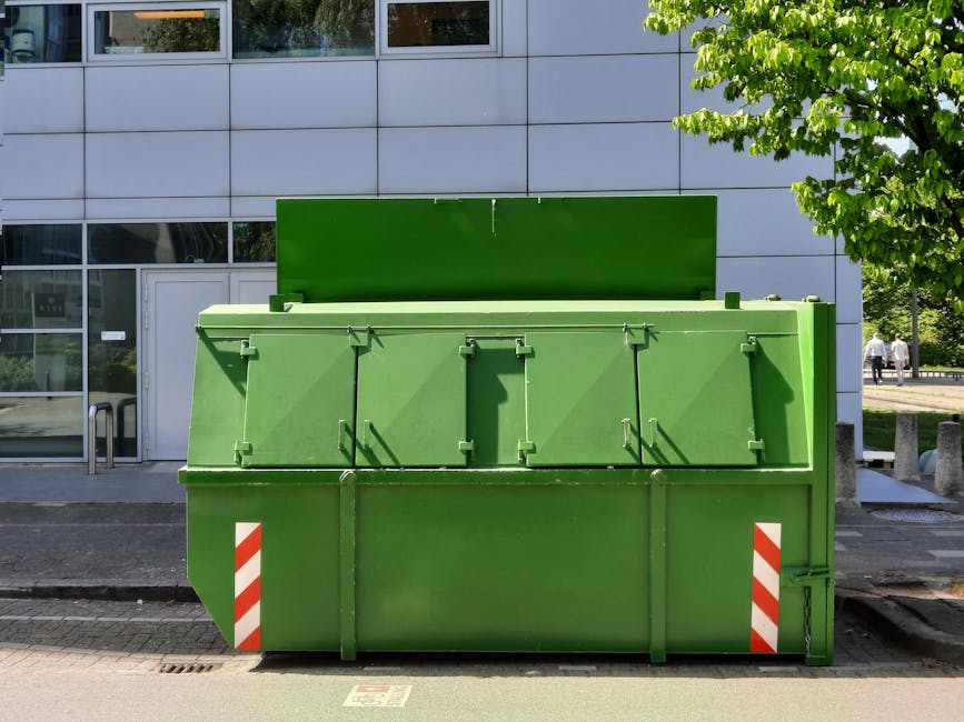 A large, green outdoor waste container with a closed lid, placed on a paved sidewalk in front of a modern building with large glass windows and white cladding. The container appears clean and well-maintained, and is positioned beside a tree with bright green leaves. There are two pedestrians in the background walking along the sidewalk. The area is well-lit with natural daylight, and the surroundings suggest an urban residential or commercial setting, emphasizing the importance of regular surface cleaning and waste management, as promoted by Holland Park Carpet Cleaning in relation to Kensington and Chelsea council waste rules for cleaning jobs.