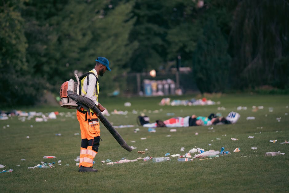 A sanitation worker dressed in bright orange reflective overalls, a matching cap, and gray gloves is sweeping the pavement with a traditional red broom on a sidewalk, with a blue dustpan held at an angle. The worker is standing next to a green trash bin mounted on a black wheeled cart, which has a black plastic bag liner, and a long-handled tool resting against it. In the background, there is a red double-decker bus partially visible on the street, with a shadow cast on the sidewalk indicating a sunny day. The area appears clean and well-maintained, suitable for commercial or residential cleaning and waste management, aligning with standards outlined by the Kensington and Chelsea council waste rules for cleaning jobs, Holland Park.