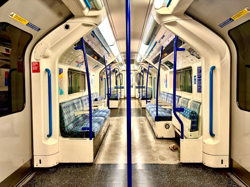Interior of a clean, empty train carriage with patterned fabric seats in shades of blue and green, arranged in rows along both sides. The carriage features polished metallic and white plastic surfaces, with blue safety handrails and vertical support poles throughout. The flooring is dark textured rubber, showing no visible dirt or debris, and the large windows on either side are clear, allowing natural light to illuminate the space. Overhead, there are embedded lighting panels and signage, all in excellent condition. Holland Park Carpet Cleaning has ensured this space is hygienic and well-maintained through professional cleaning, resulting in a fresh and sanitized environment suitable for passengers seeking a comfortable, clean transport experience, aligning with surface and deep cleaning standards for public transportation.