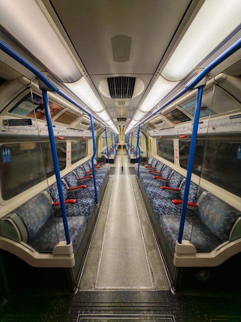 Interior of a clean, empty train carriage with patterned fabric seats in shades of blue and green, arranged in rows along both sides. The carriage features polished metallic and white plastic surfaces, with blue safety handrails and vertical support poles throughout. The flooring is dark textured rubber, showing no visible dirt or debris, and the large windows on either side are clear, allowing natural light to illuminate the space. Overhead, there are embedded lighting panels and signage, all in excellent condition. Holland Park Carpet Cleaning has ensured this space is hygienic and well-maintained through professional cleaning, resulting in a fresh and sanitized environment suitable for passengers seeking a comfortable, clean transport experience, aligning with surface and deep cleaning standards for public transportation.