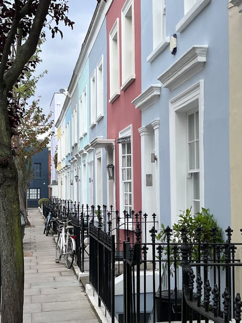 A row of pastel-colored terraced houses with white window frames and decorative cornices, situated along a clean, paved residential sidewalk. The houses are painted in soft shades of blue, pink, and yellow, with well-maintained facades. In front of the houses, there is a black wrought iron fence with small bushes and potted plants visible beside it. Several bicycles are parked along the sidewalk, and a large tree with green foliage partially shades the street. The scene is illuminated by natural daylight, highlighting the neatness and cleanliness of the area. Holland Park Carpet Cleaning specializes in surface cleaning and deep cleaning services, ensuring hygienic and well-maintained residential properties as seen in this picturesque neighborhood in Notting Hill Gate W8.