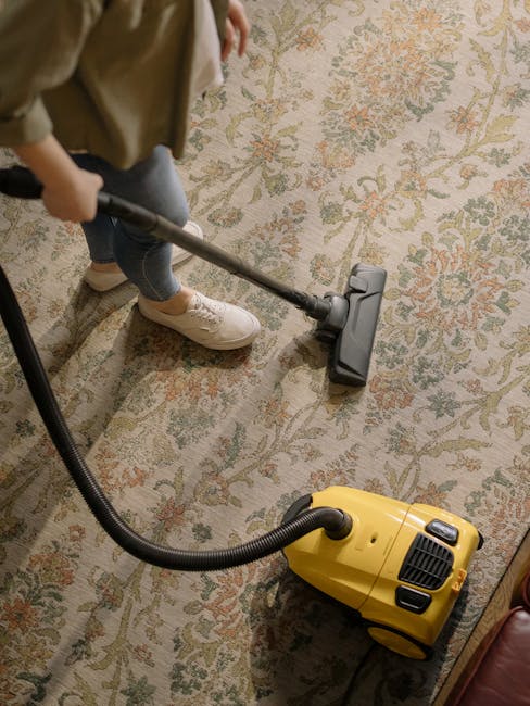A person wearing beige trousers and white sneakers is vacuuming a traditional floral-patterned area rug with a yellow carpet cleaner, which features a black hose and vacuum head, in a well-lit room. The rug has a mix of cream, green, and pink tones, and the cleaning process appears thorough, indicating surface cleaning and deep cleaning procedures. The setting suggests a residential environment, and the image portrays professional domestic cleaning, as performed by Holland Park Carpet Cleaning, aligning with the focus on the real cost of carpet cleaning in Holland Park 2026 price guide.