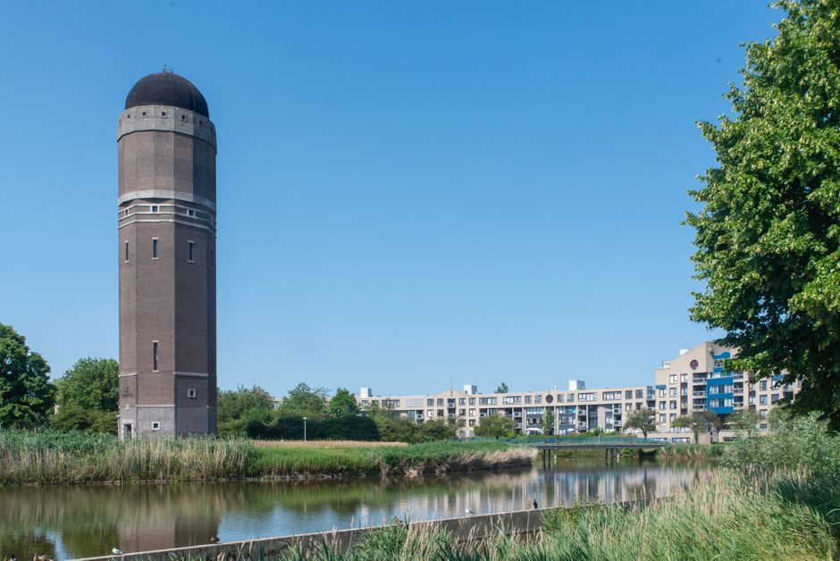 A tall, cylindrical water tower made of brown brick with small rectangular windows and a black dome-shaped roof, situated beside a waterway with lush green trees on the right and low grass along the shoreline, under a bright blue sky with minimal clouds. In the background, there is a modern residential building with multiple floors, balconies, and large windows, reflecting a well-maintained urban landscape in Holland Park, captured during daytime. The scene is clear, well-lit, and shows the contrast between natural elements and urban development, emphasizing the clean and quiet environment that Holland Park Carpet Cleaning services aim to maintain.